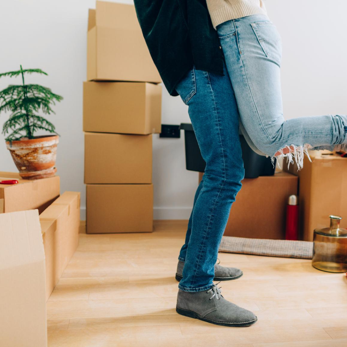 Symbolbild für einen erfolgreichen Immobilienverkauf und Einzug: Ein Paar umarmt sich freudig in seinem neuen Zuhause, wobei eine Person die andere anhebt. Um sie herum stehen gestapelte Umzugskartons auf einem hellen Holzboden, ergänzt durch eine Zimmerpflanze. Ein emotionales Motiv, das den Traum vom Eigenheim, den erfolgreichen Hauskauf und den Start in einen neuen Lebensabschnitt perfekt visualisiert.
